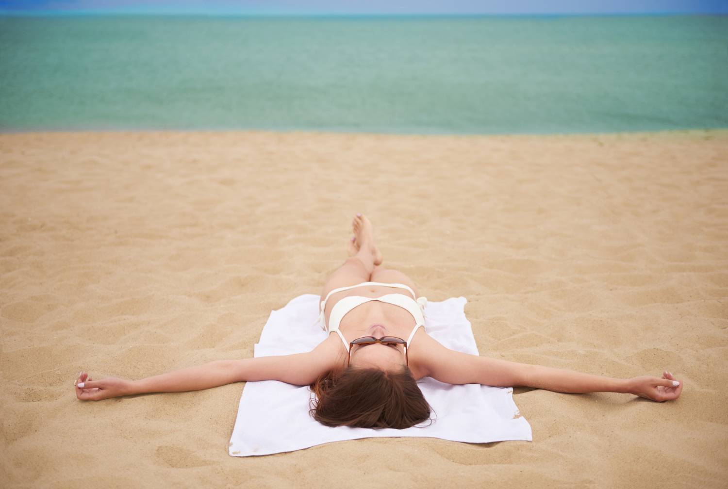 A woman in a white bikini relaxes on a towel on a sandy beach, with the serene turquoise ocean stretching out in the background.