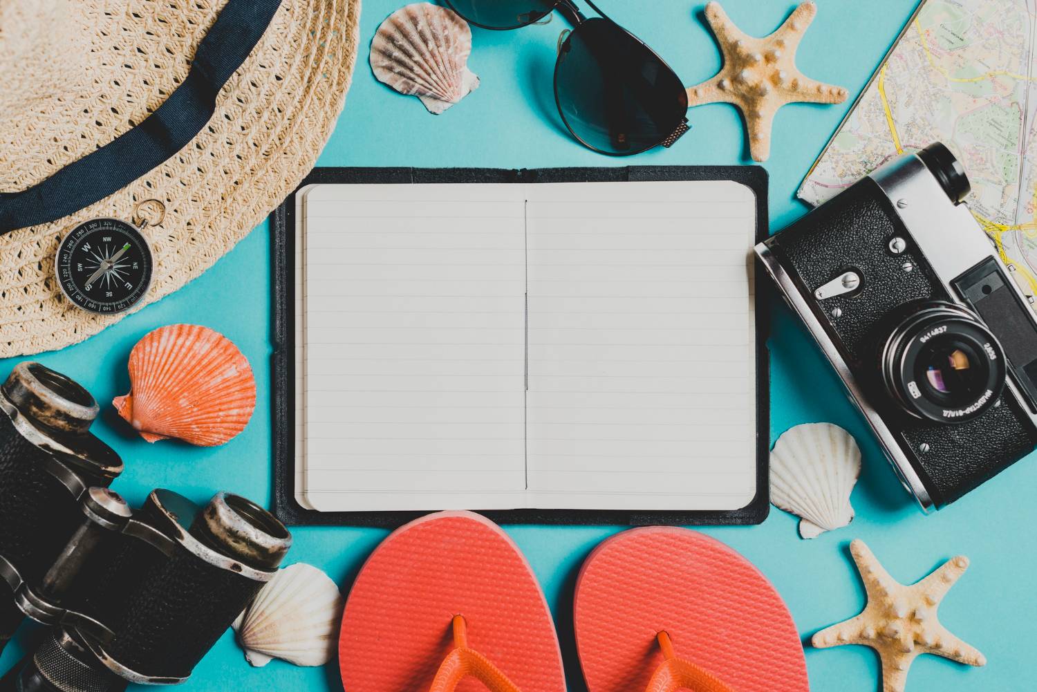 A summer travel setup featuring a straw hat, compass, camera, sunglasses, flip-flops, seashells, starfish, and an open notebook on a blue background.
